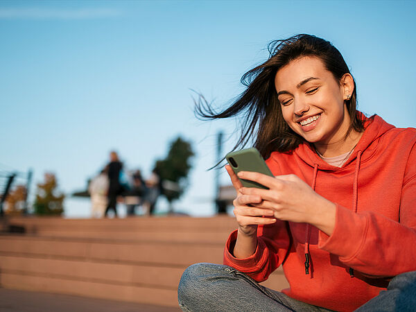 Junge Frau in rotem Hoodie sitzt draußen und schaut lächelnd auf ihr Smartphone; im Hintergrund sind unscharf Menschen und eine Treppe zu sehen.