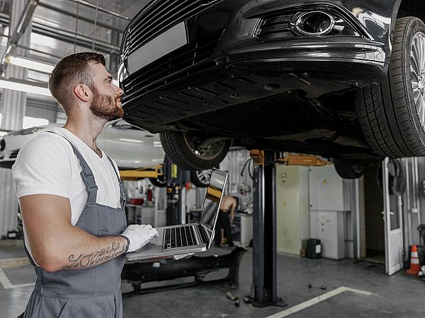 Ein Mechaniker steht mit dem Laptop vor einer Autohebebühne und sieht sich das Auto in der Werkstatt an.