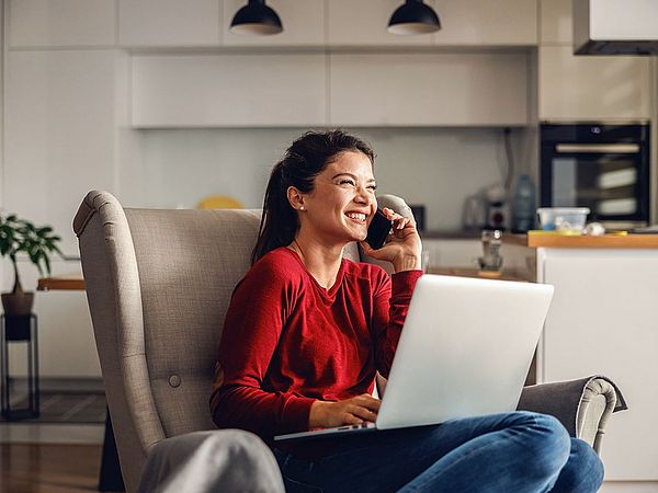 Eine glückliche Frau sitzt mit ihrem Laptop auf einem Sofa im Wohnzimmer und telefoniert