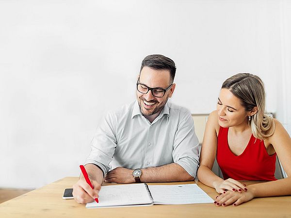 Ein lächelnder Mann und eine Frau sitzen gemeinsam am Schreibtisch, wobei Notizen auf einem Zettel gemacht werden. 